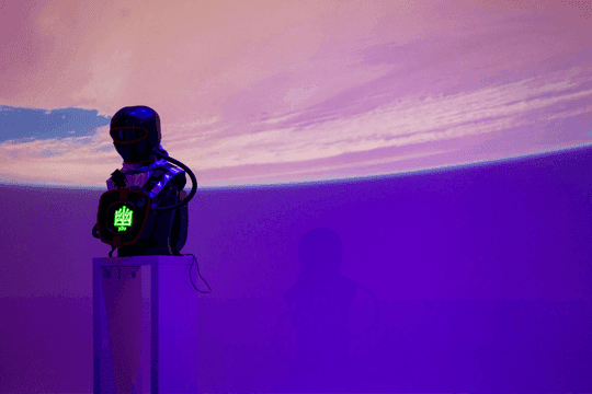 Front detail of the astronaut-like Hidden sculpture, chest-piece display glowing green, against a pink-and-purple projected sky.