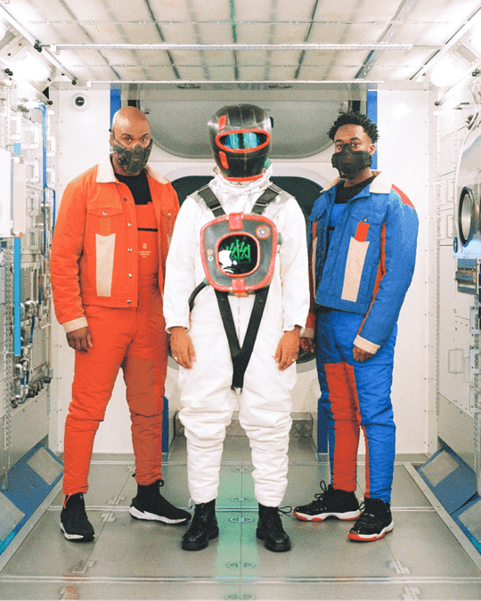 Campaign portrait: two models in orange and blue Secluded jackets flanking the Hidden sculpture in its full astronaut suit.