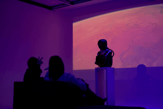 Hidden on its pedestal against a projected planetary sky, silhouetted visitors seated in front of it at the release party.