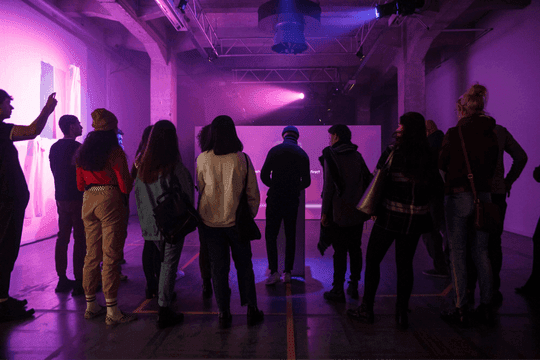 The industrial hall installation: a crowd gathered under purple gallery lighting to watch the projection.