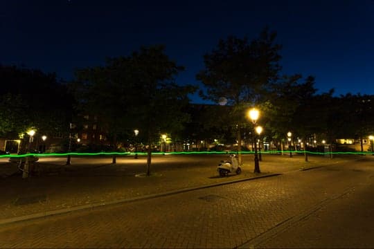 A night shot in an urban plaza: the green trail runs between lamppost pools of orange light.