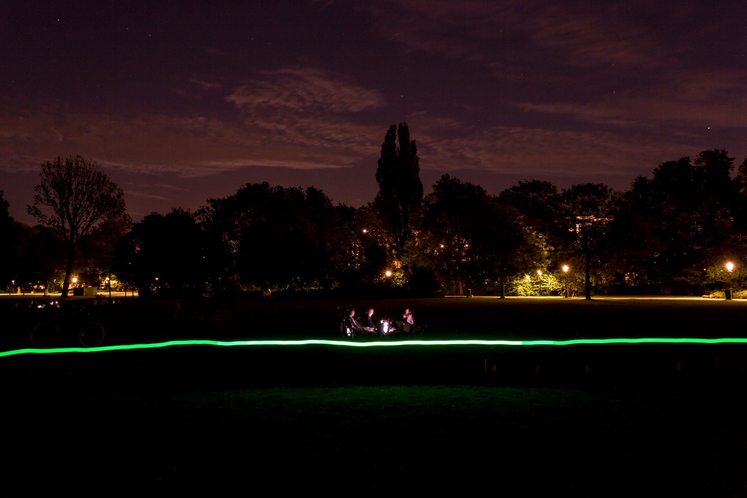 Long-exposure photograph of a WiFi network's signal strength, traced as a wavering line of green light across a dark Amsterdam park, three figures gathered where the signal peaks.
